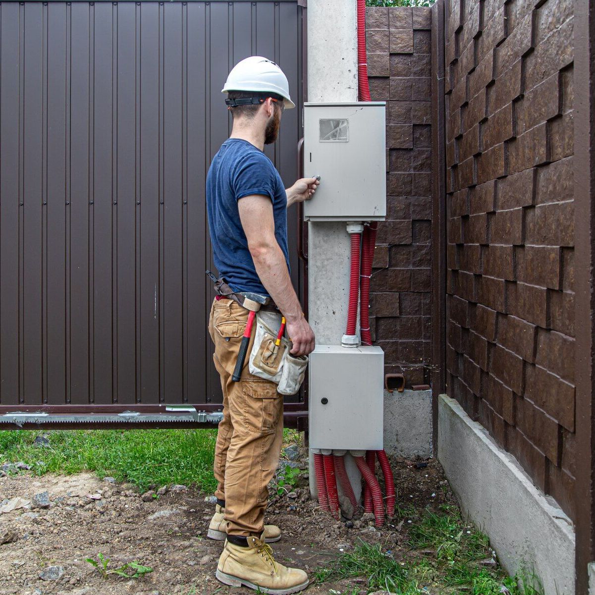 Worker in hard hat inspecting an outdoor electrical panel beside a brick wall