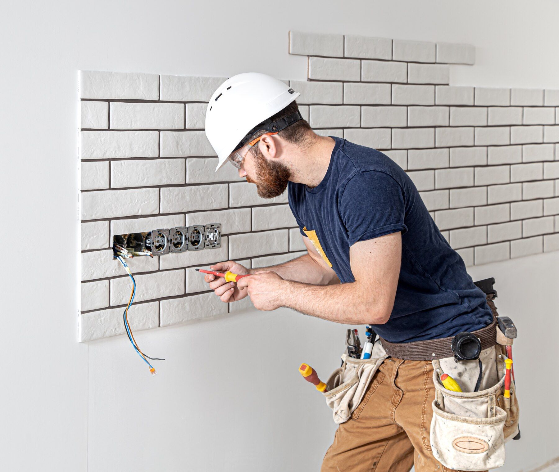 Electrician in a white hard hat wiring a wall outlet opening with exposed cables and tiled backsplash.