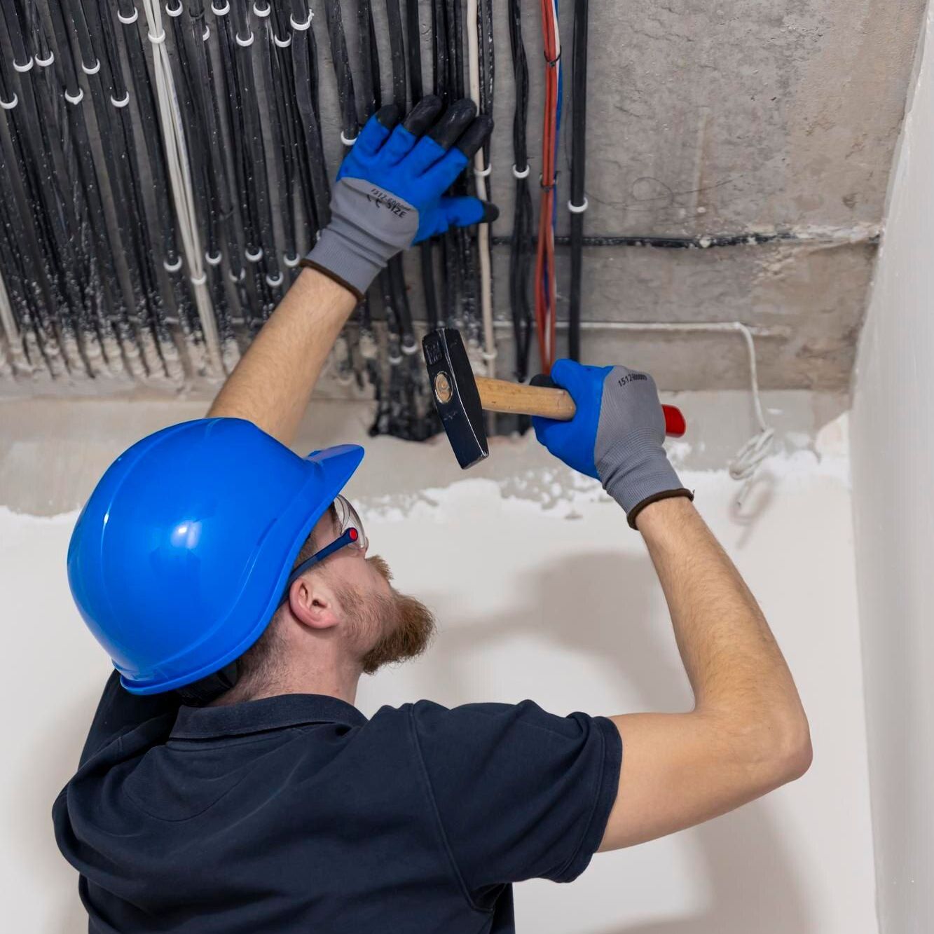 Worker in blue hard hat inspects ceiling wiring with tools at an unfinished construction site