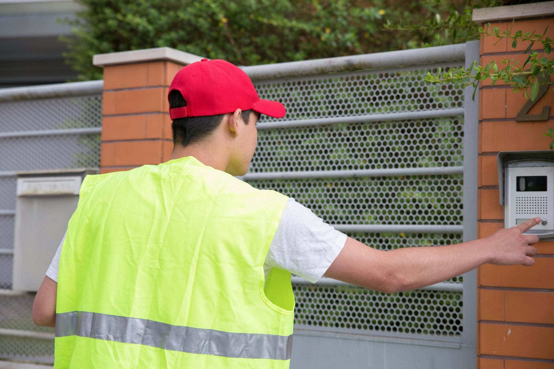 Man in vest and cap rings a doorbell on a gated property.