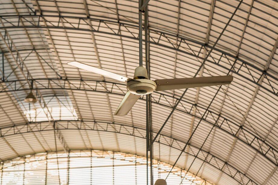 Large ceiling fan under an arched metal roof in a bright indoor hall