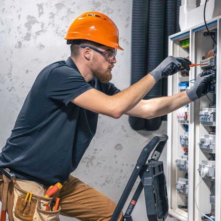 Technician in orange hard hat wiring an electrical panel on a ladder