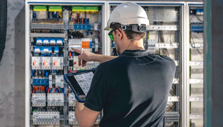 Technician in a hard hat inspecting an open electrical control panel with a tablet.