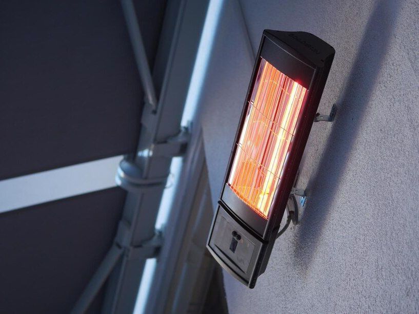 Wall-mounted outdoor heater glowing orange beside a gray wall and metal railing.