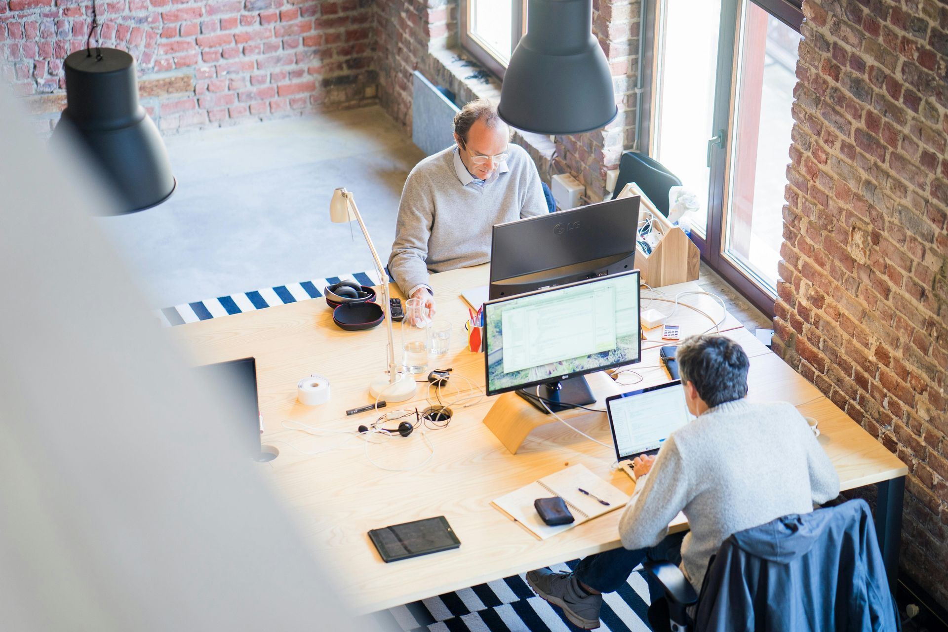 Two men work at a desk in a modern office with exposed brick, using computers.