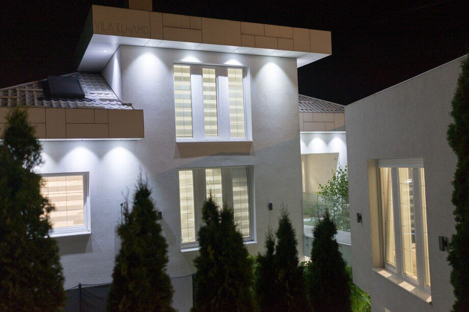 Modern white house exterior at night with bright porch lights and tall shrubs in front.