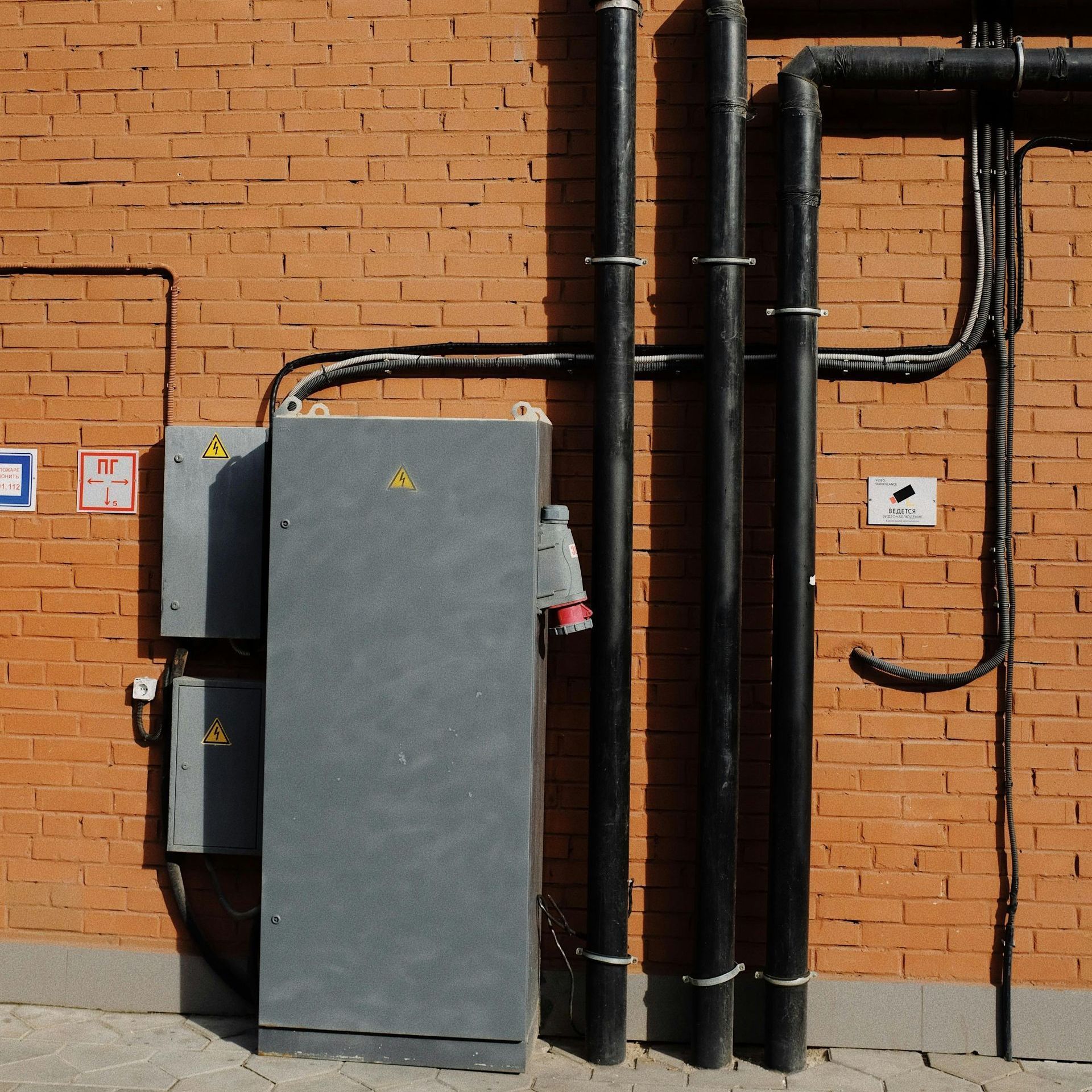 Electrical boxes and pipes on a brick wall. Gray boxes with warning signs, black pipes, and red brick.