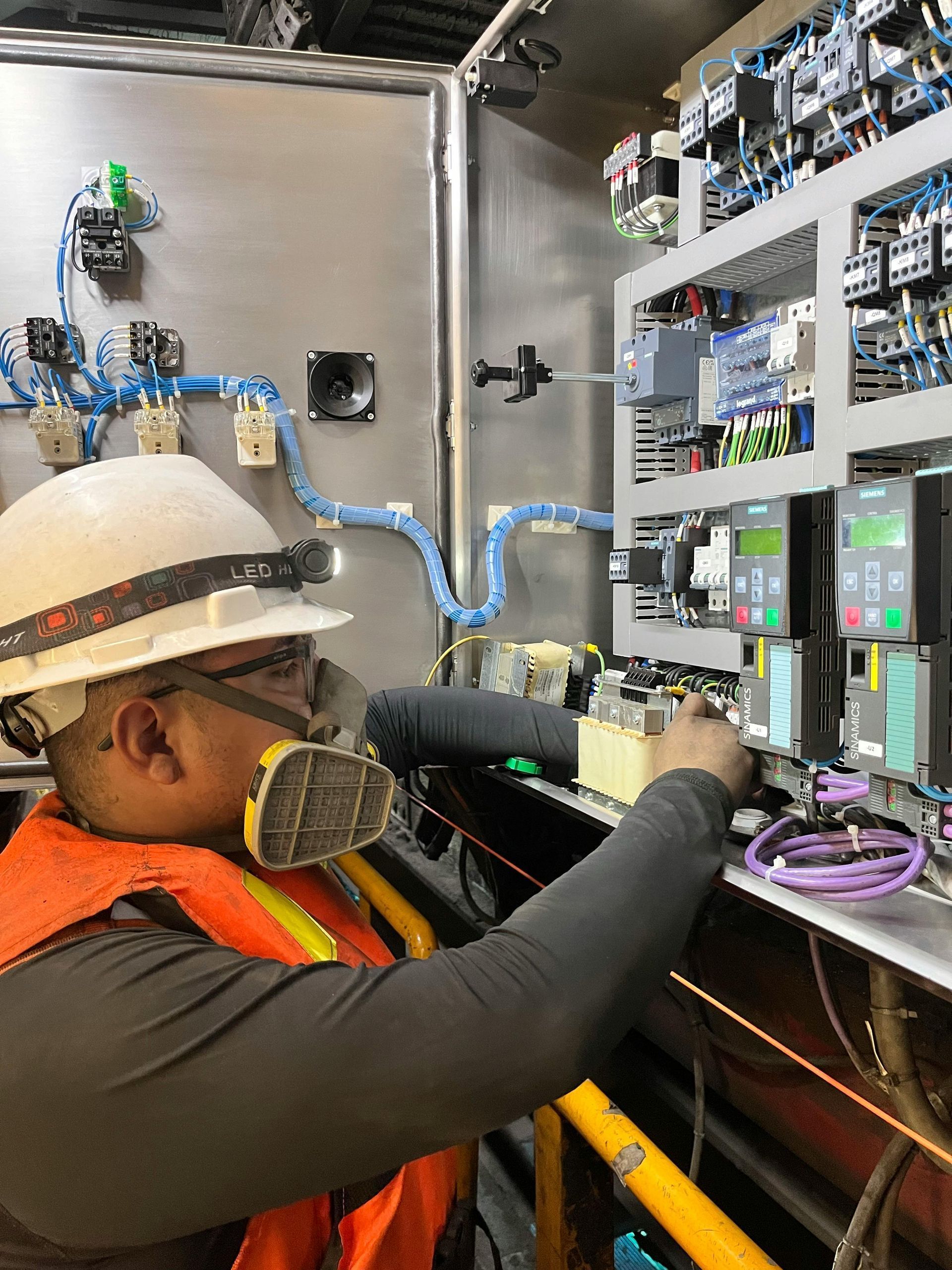 Electrician in hard hat and respirator working inside an electrical panel, wearing safety vest.