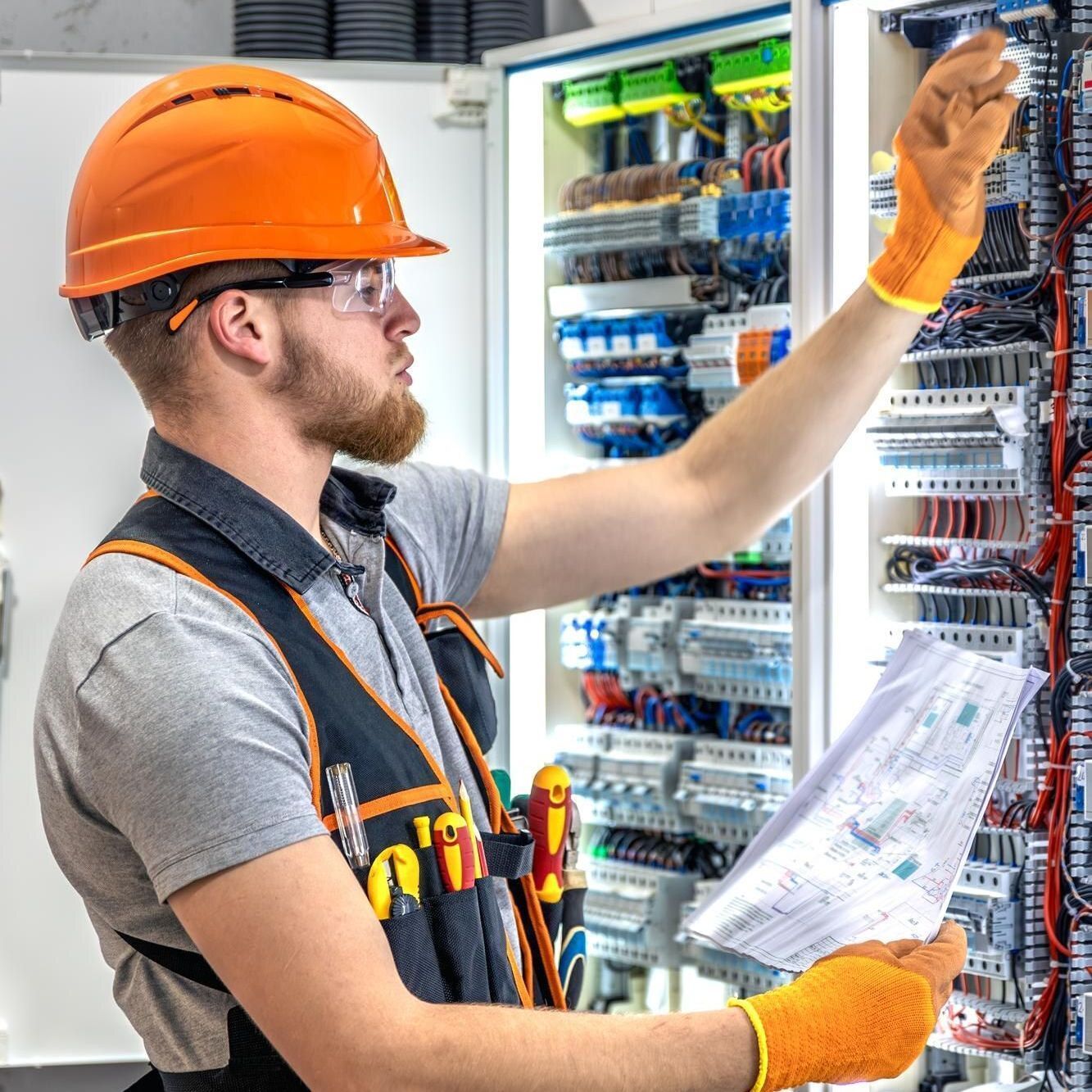 Electrician in an orange hard hat and gloves inspecting a circuit panel, holding a blueprint.