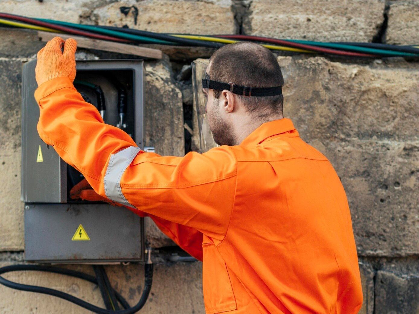Worker in orange safety jacket inspecting an outdoor electrical box on a stone wall