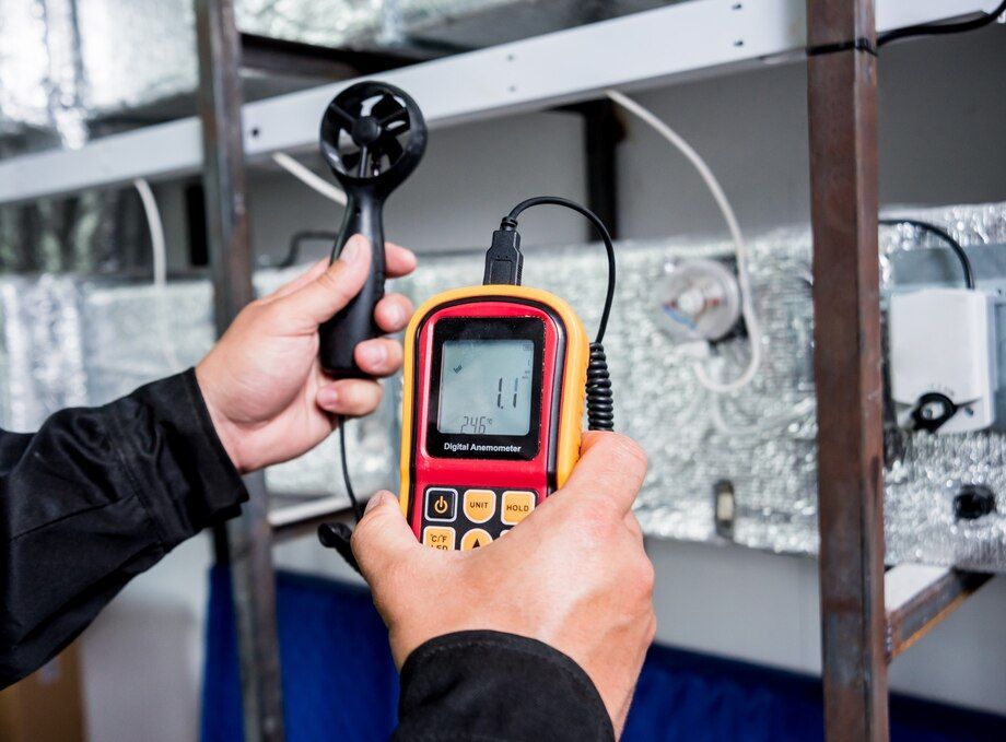 Hands using a red handheld meter and black probe to inspect HVAC equipment indoors