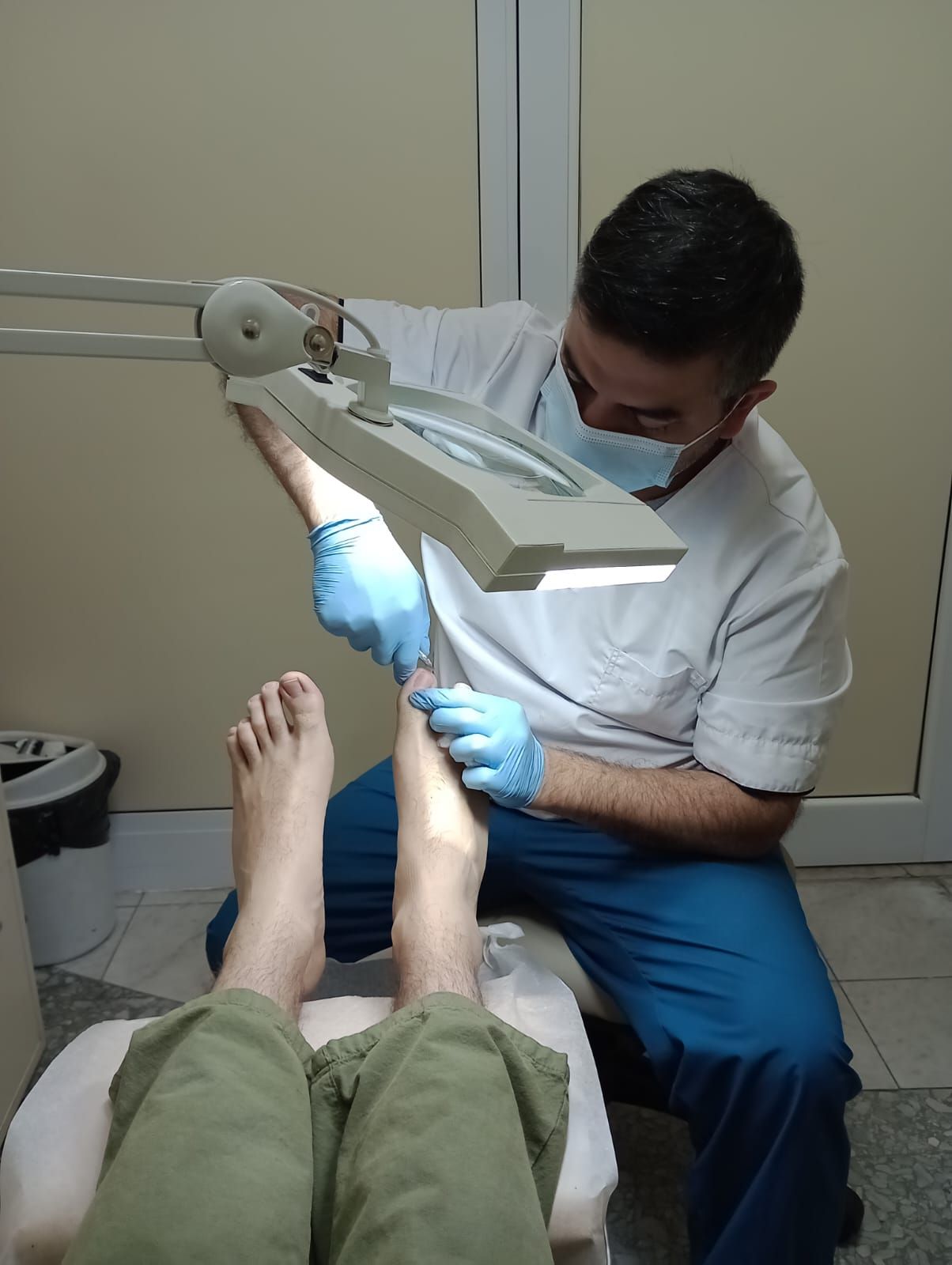 Manos con guantes azules usando un bisturí en la planta del pie. Otra persona al fondo.