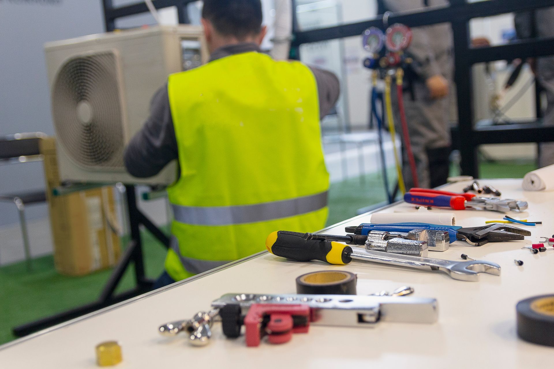 Tools on a workbench with a technician servicing an HVAC unit in the background. Tools on a workbench with a technician servicing an HVAC unit in the background.
