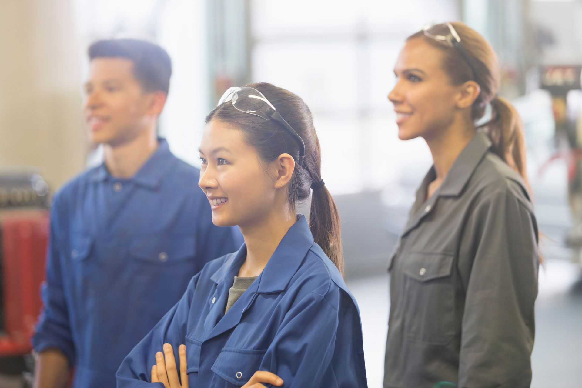 Three trainees in safety glasses listening during hands-on automotive workshop training. Three trainees in safety glasses listening during hands-on automotive workshop training.