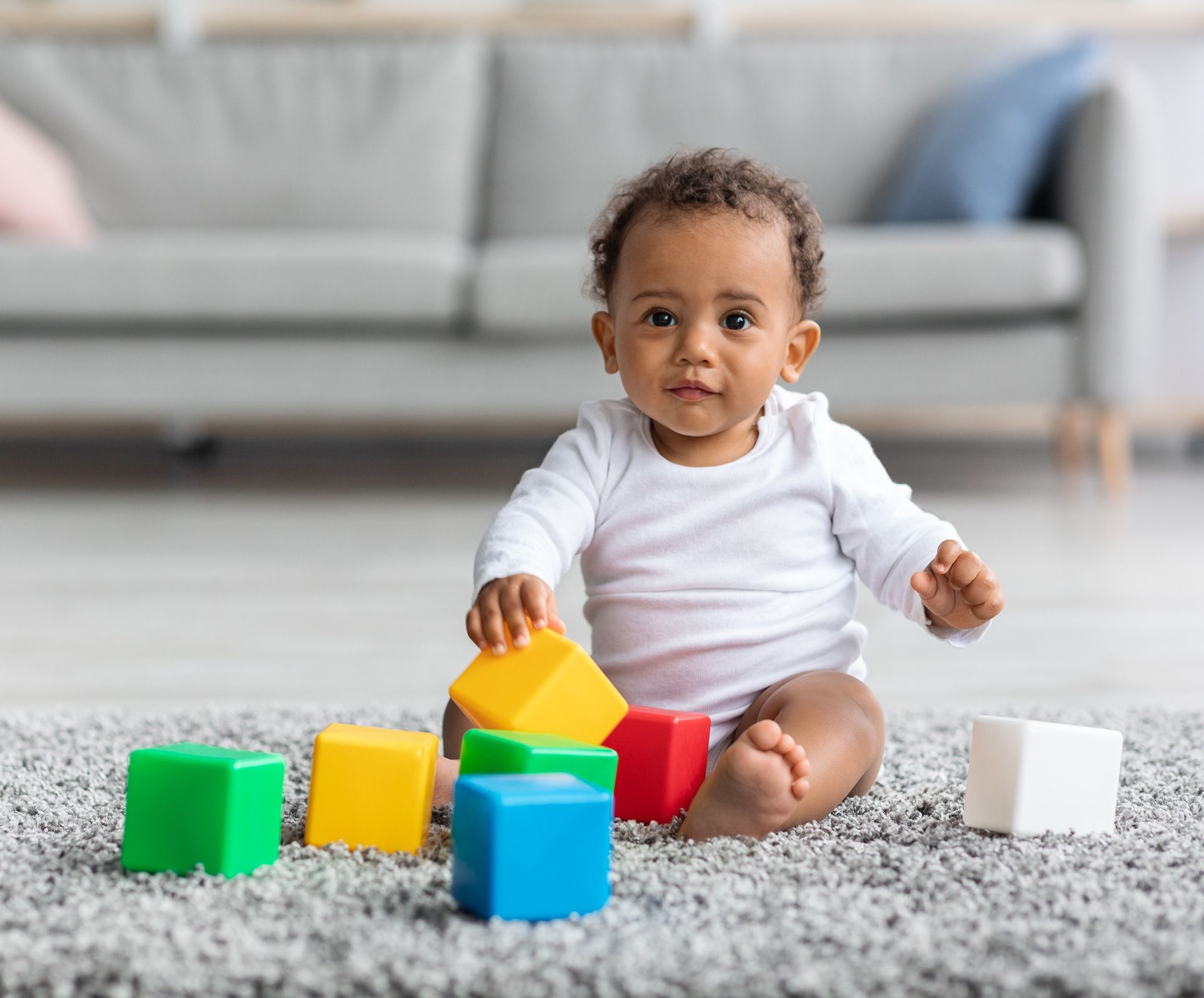 A baby in a white shirt plays with colorful blocks on a grey carpeted floor in a living room.