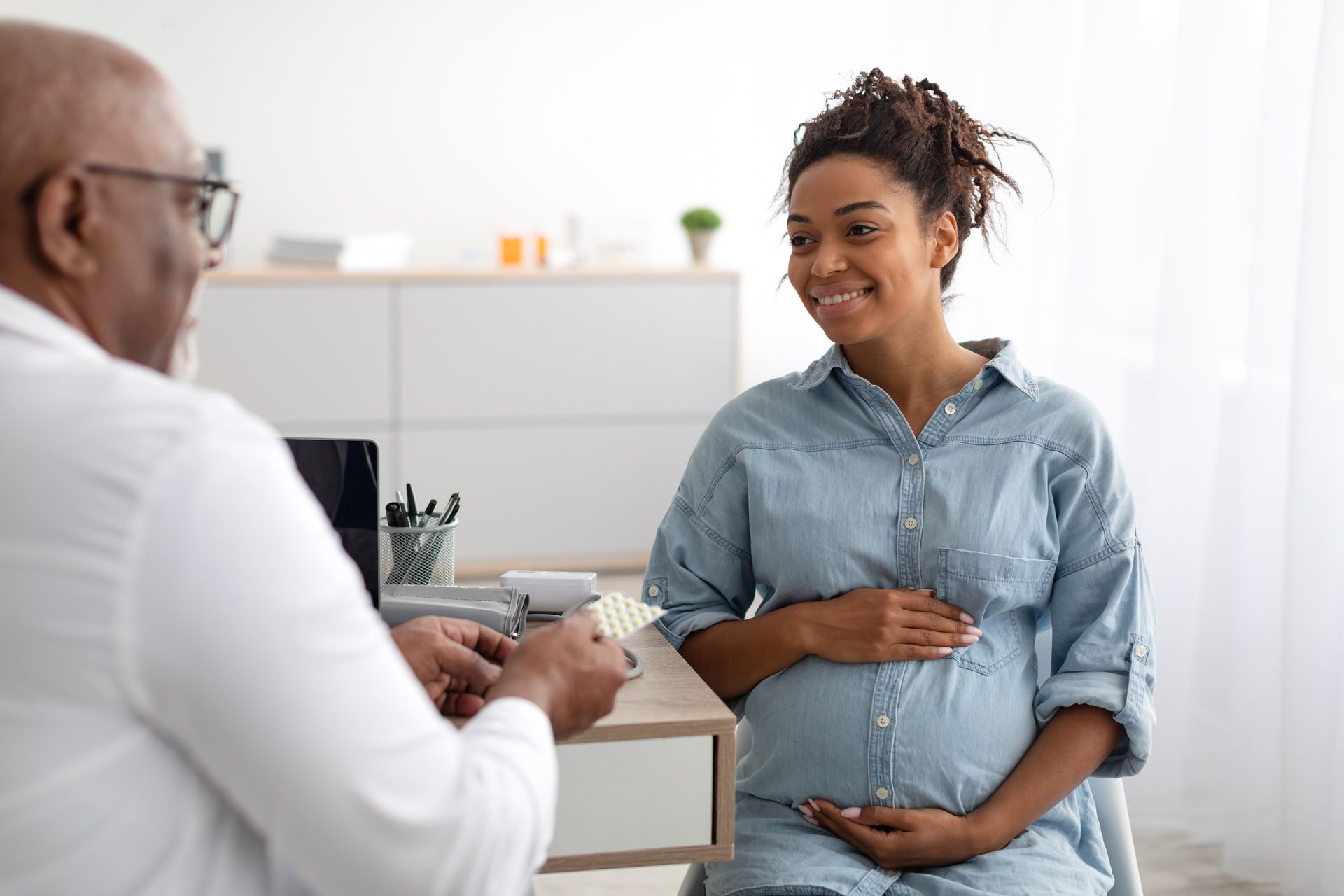 A healthcare professional hands a packet of pills to a smiling expectant person in a bright office.