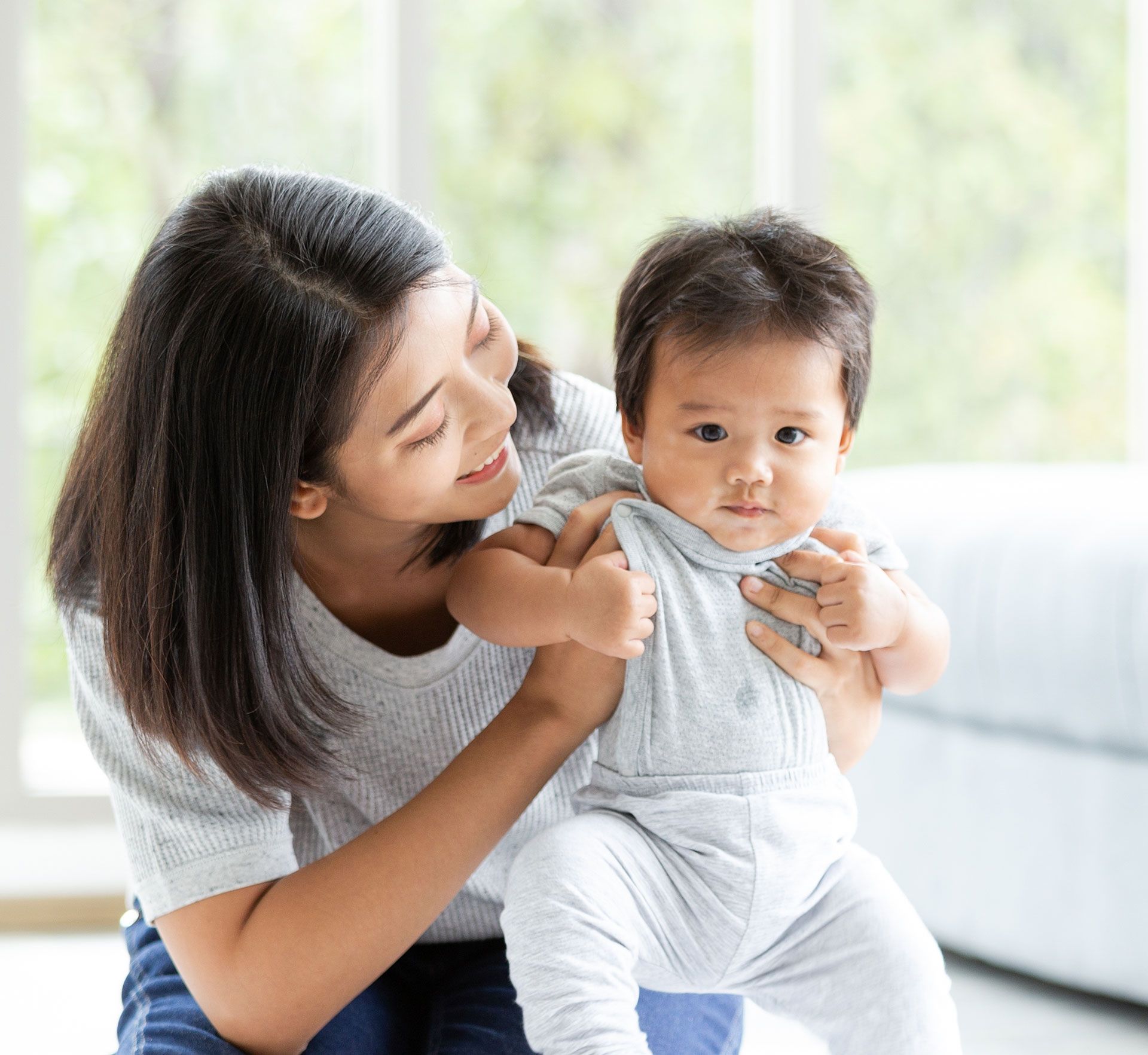 A person in a light gray shirt smiles while holding a baby who is wearing a matching light gray onesie.