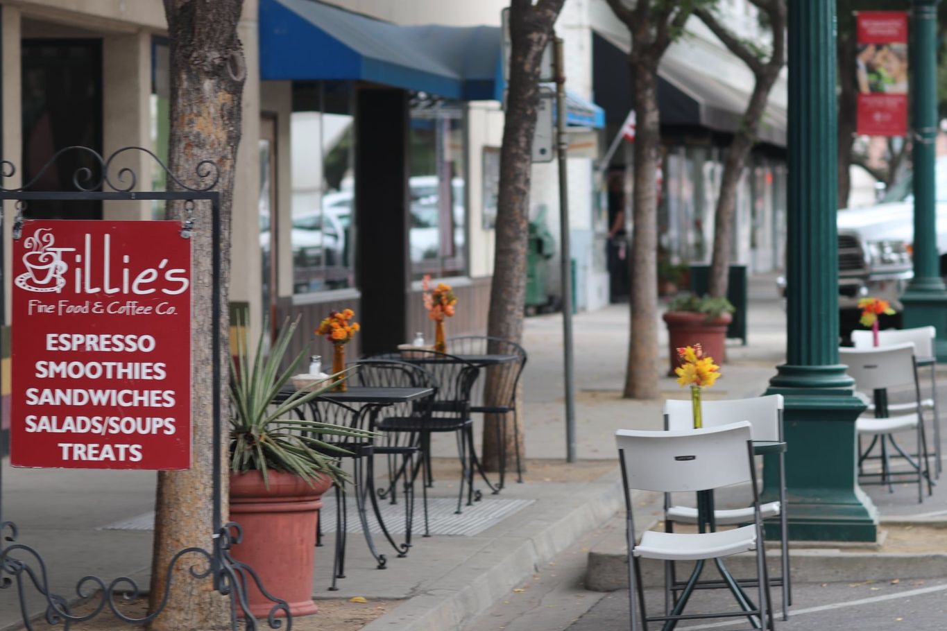 Sidewalk café with red menu sign, empty tables, flowers, and storefronts along a tree-lined street.