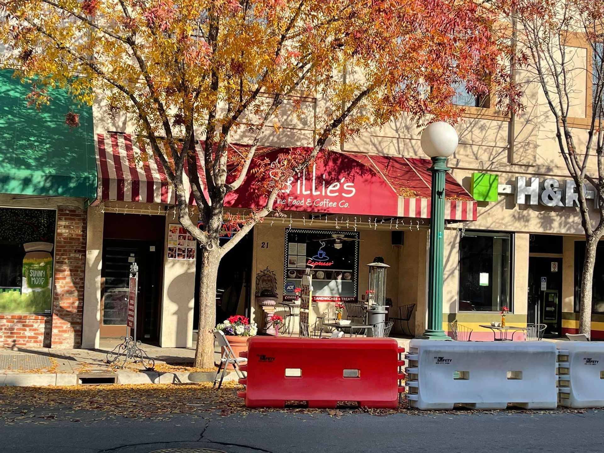 Small storefronts under autumn trees, with red and white barricades in front.