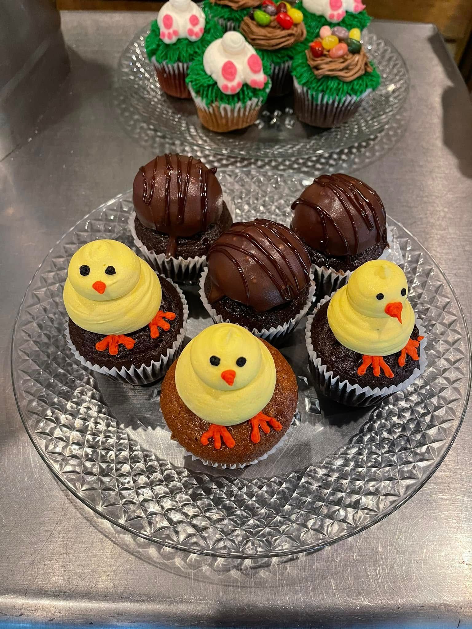 Cupcakes on glass platters, decorated like chicks, green eggs, and chocolate drizzles on a metal counter