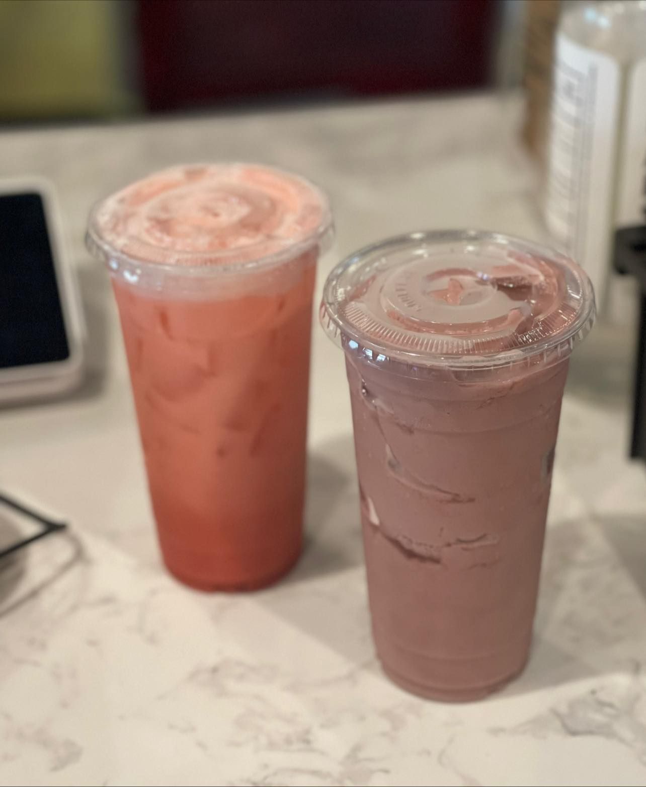 Two iced drinks in clear cups on a marble countertop, one coral pink and one pale lavender.
