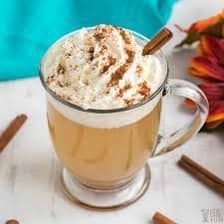 Glass mug of coffee topped with whipped cream and cinnamon on a white table, with cinnamon sticks nearby