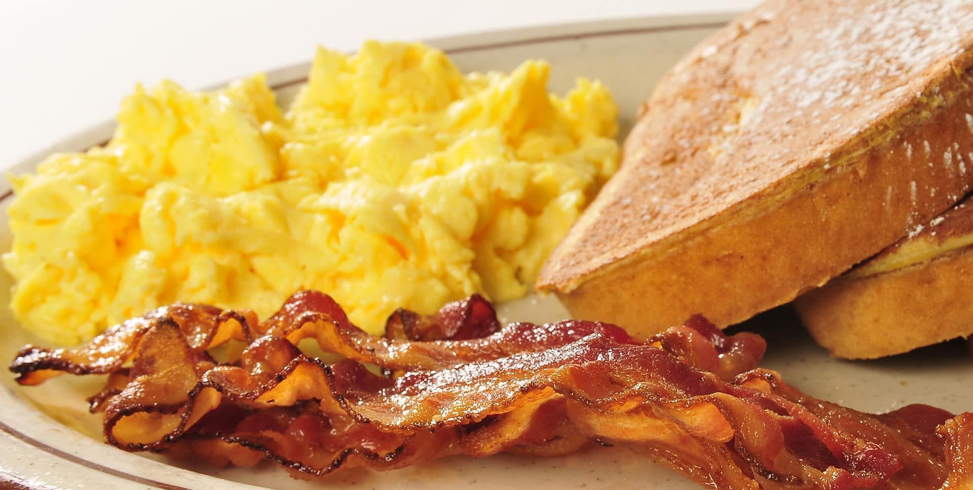 Scrambled eggs, bacon, and toast on a plate, with a breakfast sandwich in the background