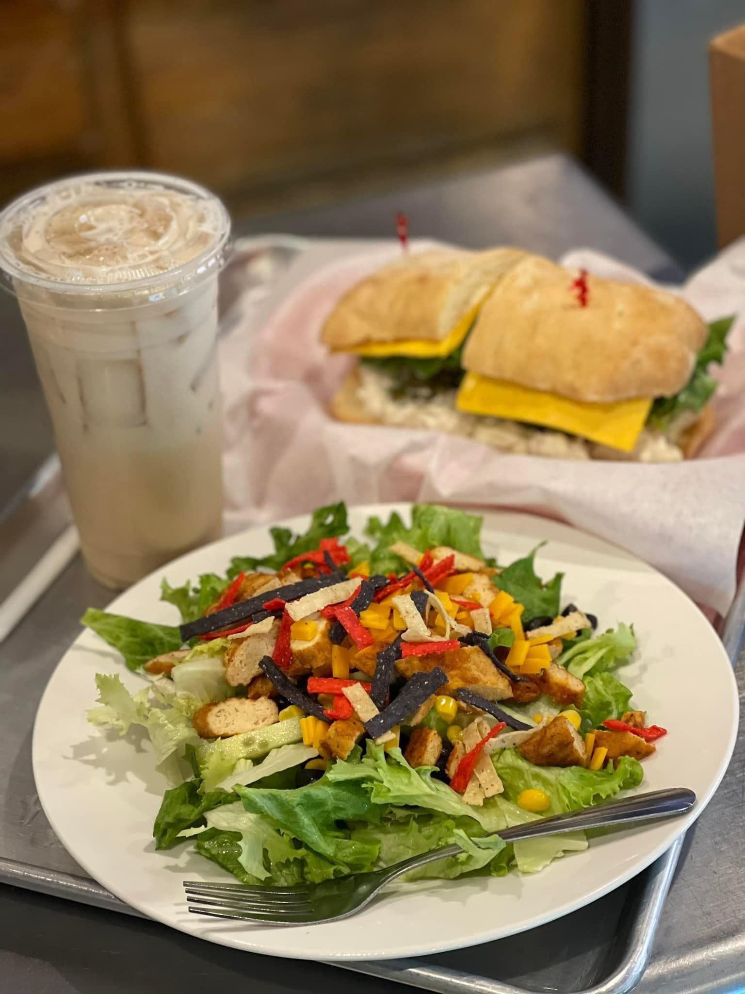 Salad plate and iced drink in foreground, with a sandwich on a tray behind them.
