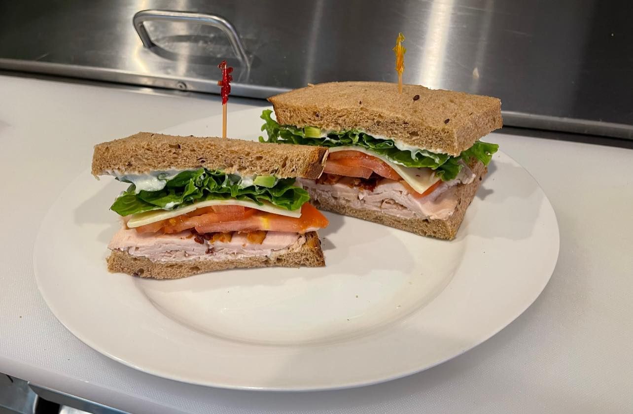 Two club sandwiches on a white plate in a kitchen, with lettuce, tomato, and deli meat visible