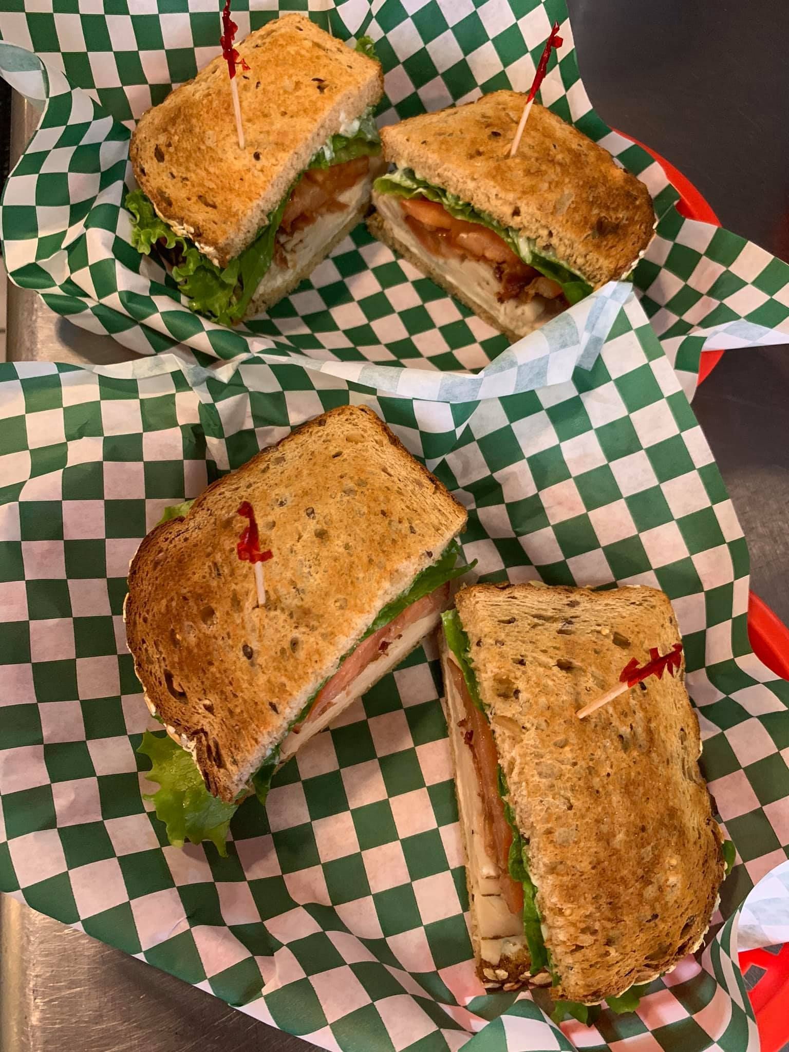 Four sandwich halves on green-and-white checkered paper in a basket, with lettuce and visible fillings.