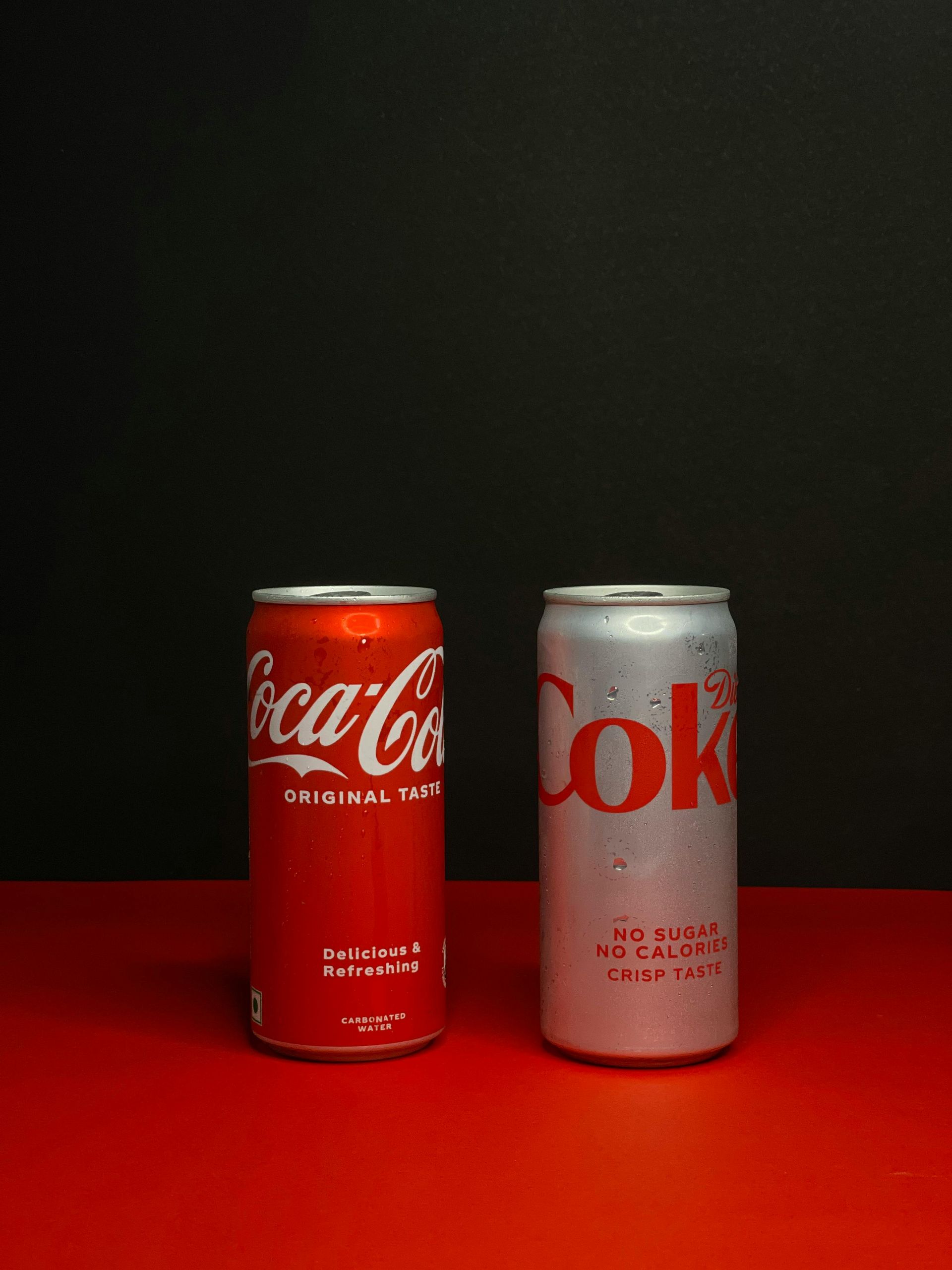Two Coca-Cola cans, one red and one silver, standing side by side on a red surface against a black background.