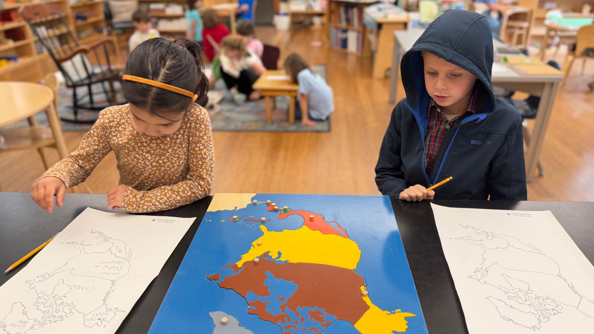 Image of two elementary children sitting at a table with a puzzle map of North America between them, and drawing paper to copy it