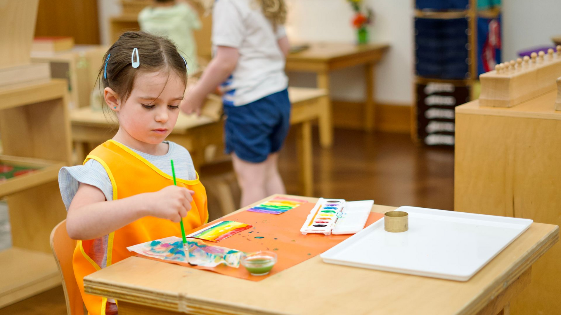 Image of a preschool aged girl sitting at a small table with a palette of water colors and a few sheets of paper