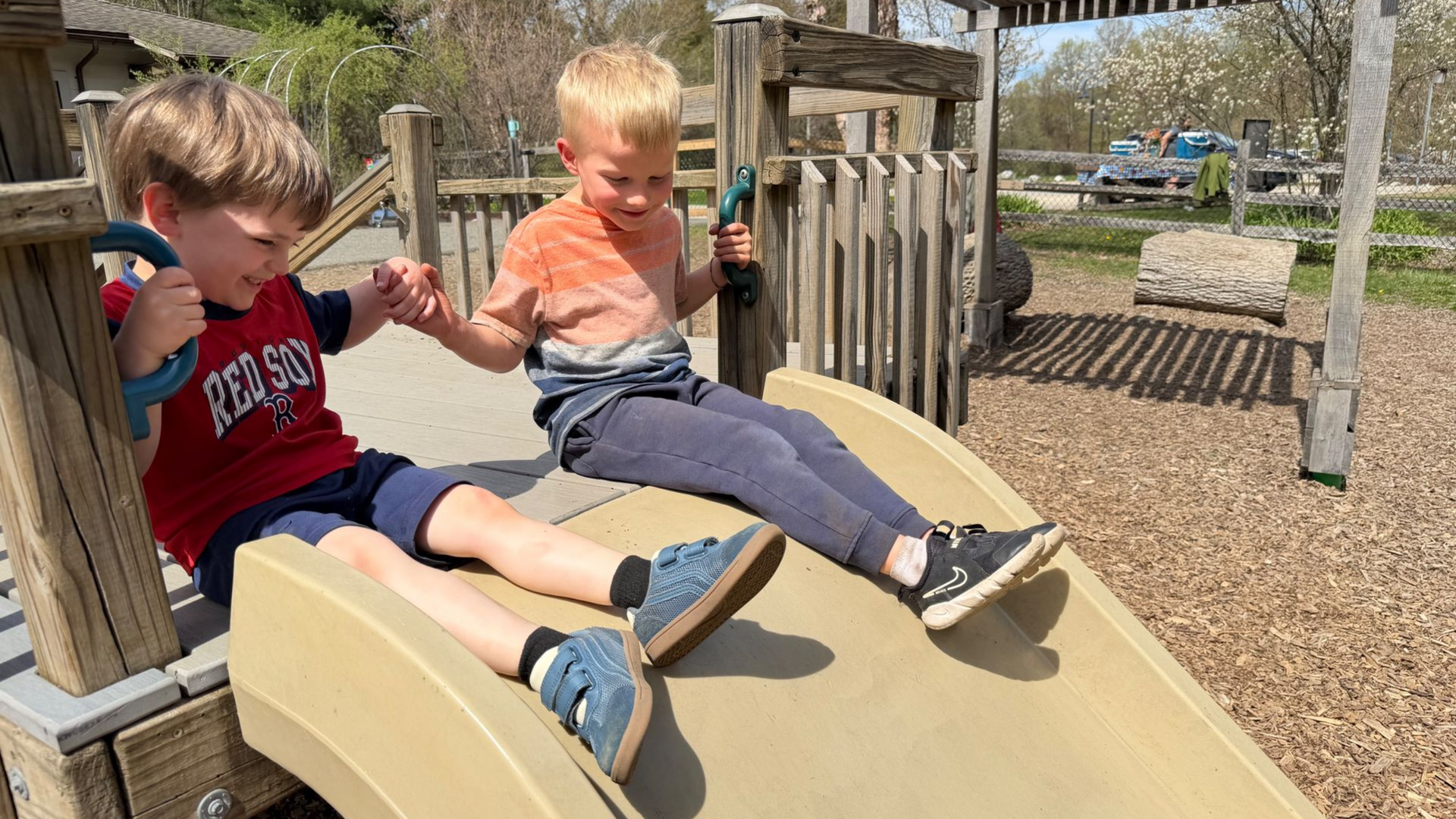 two preschool aged children sitting at the top of a slide, smiling and holding hands
