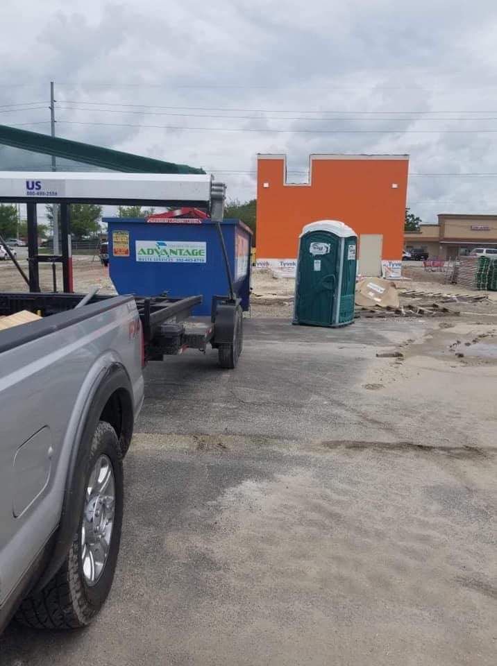 A truck is towing a dumpster and a portable toilet in a parking lot.