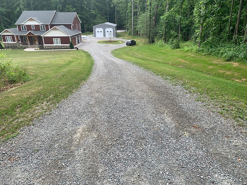 A gravel driveway leads to a large, multi-story house and a separate two-car garage set against a wooded landscape.