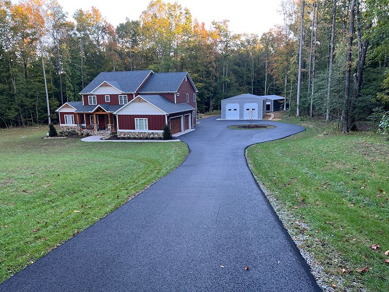A large red suburban home with a long asphalt driveway leading to a detached two-car garage, surrounded by forest.