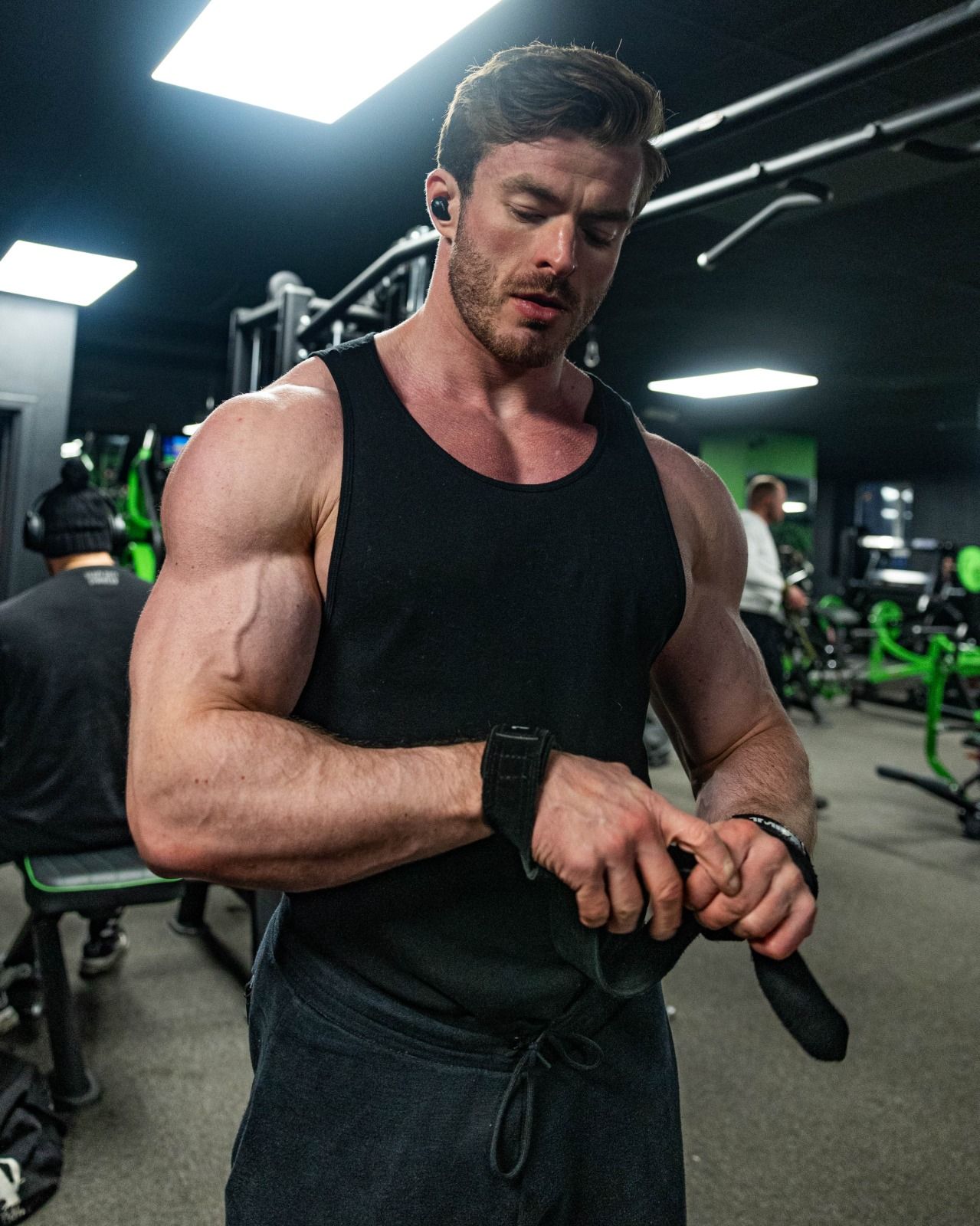 Muscular man in a black tank top adjusting wrist straps in a gym with green equipment