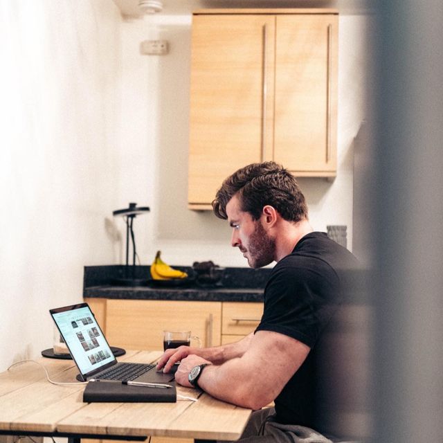 Man working on a laptop at a kitchen table with a black keyboard and banana on the counter behind him