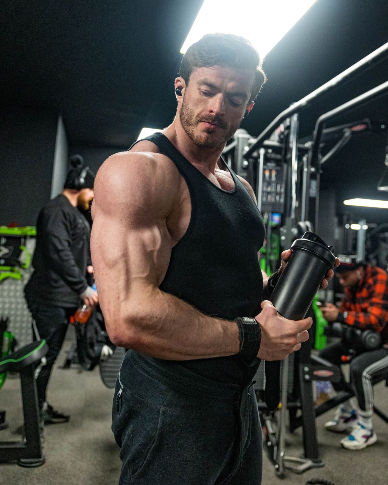 Man flexing bicep in a gym, wearing a black tank top and holding a water bottle