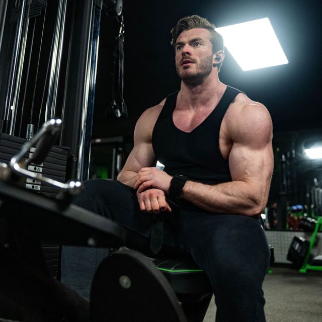Muscular man in a black tank top sitting in a gym, with workout equipment and bright overhead lights.