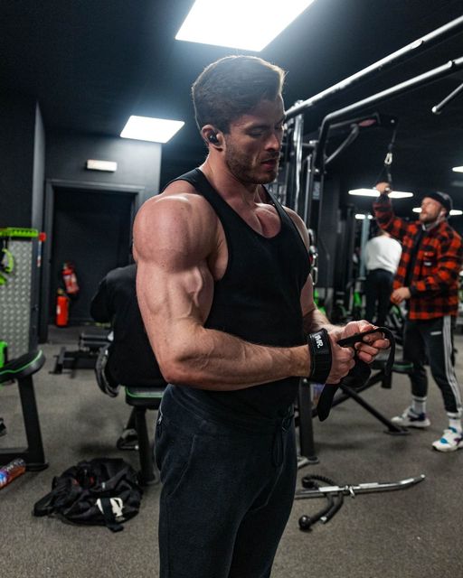 Man in black tank top flexing in a gym, with cable machines and another person in the background