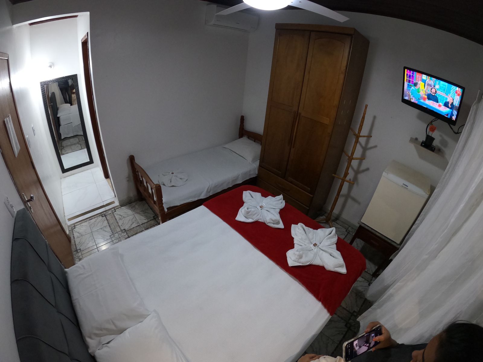 Quarto de hotel com duas camas, guarda-roupa, TV e porta aberta para o corredor com espelho.