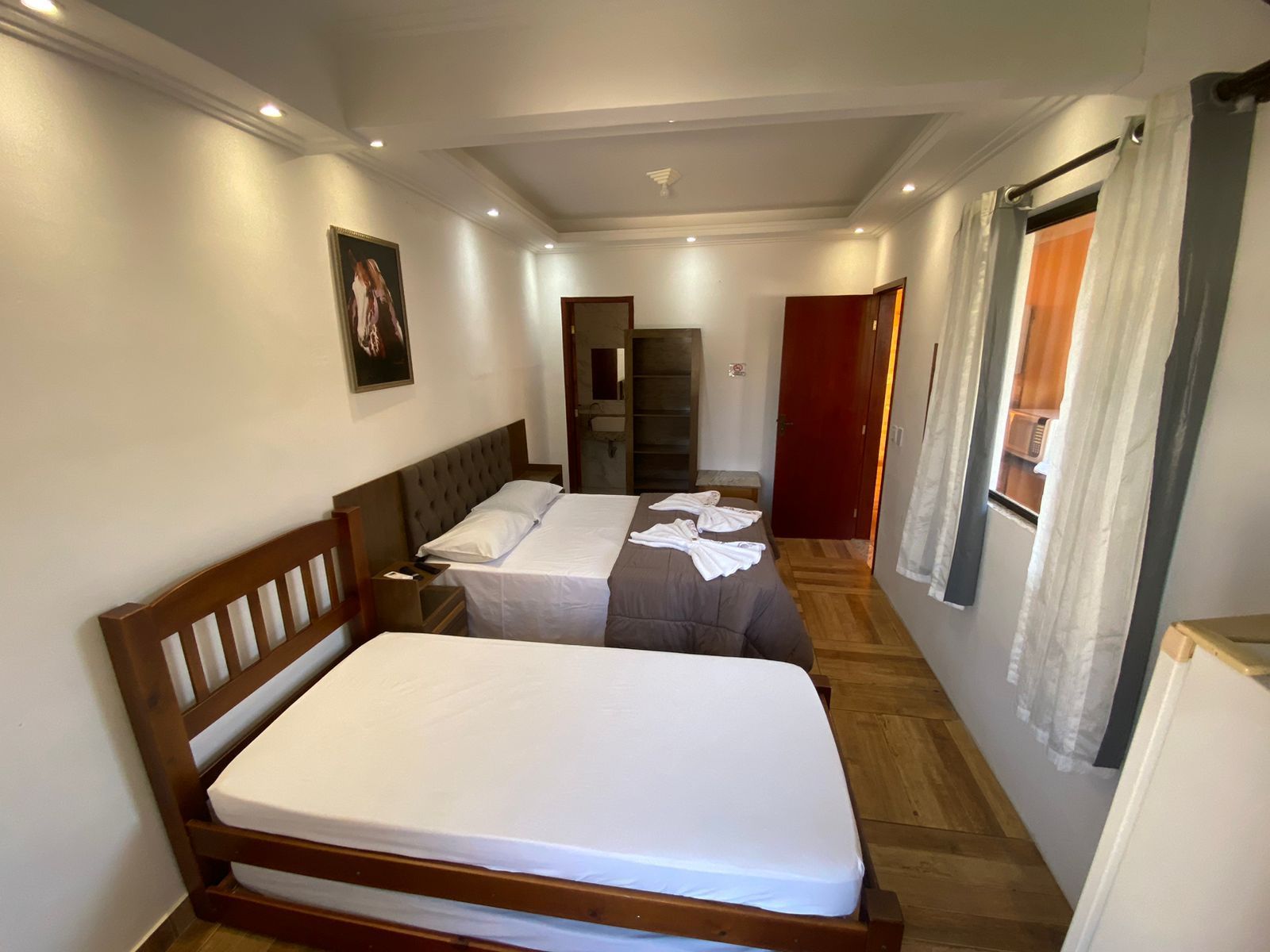 Quarto com duas camas, lençóis brancos, estrutura de madeira, porta para o banheiro e entrada.