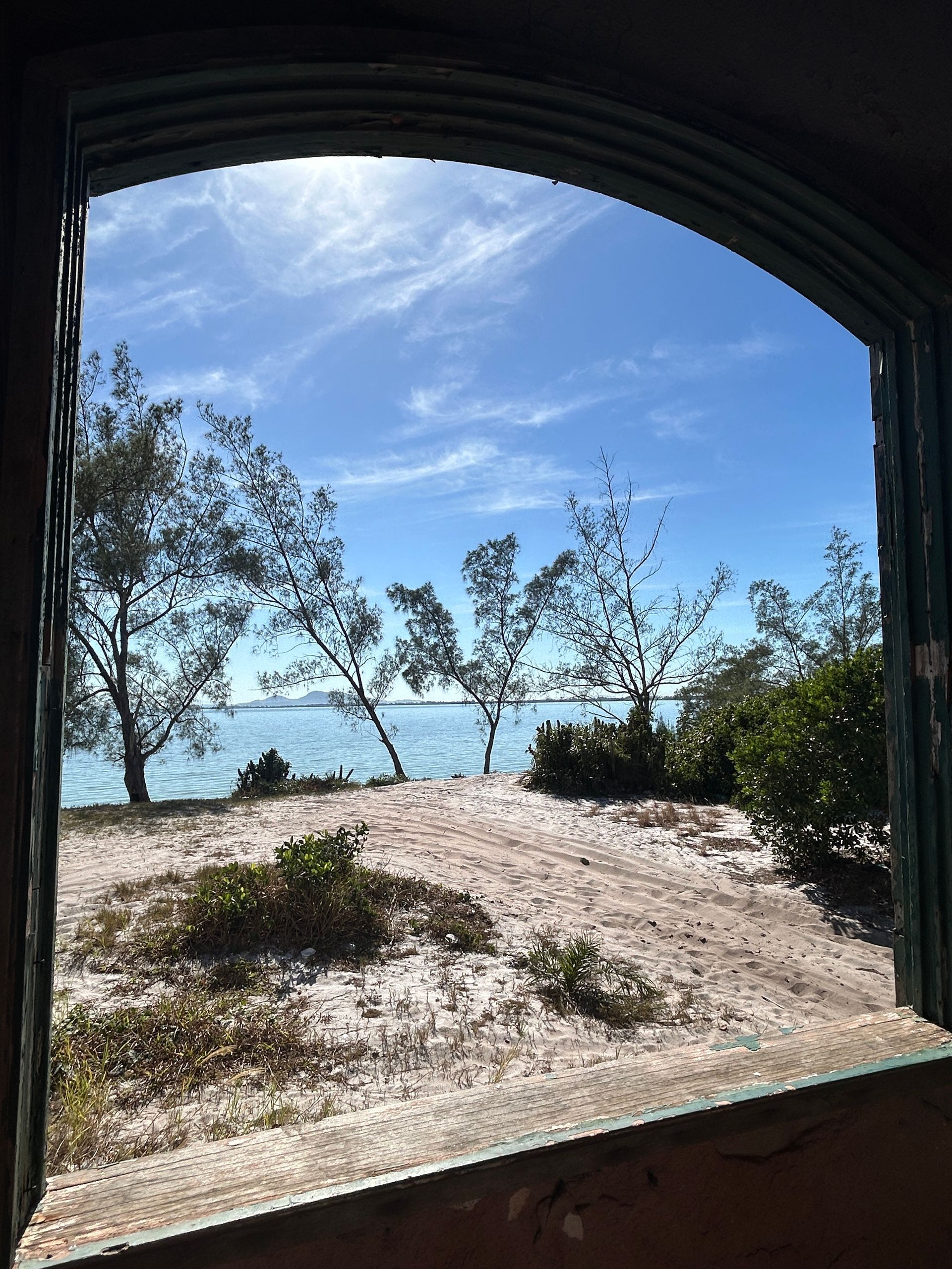 Vista de um arco desgastado pelo tempo para uma costa arenosa com árvores e céu azul sobre a água.