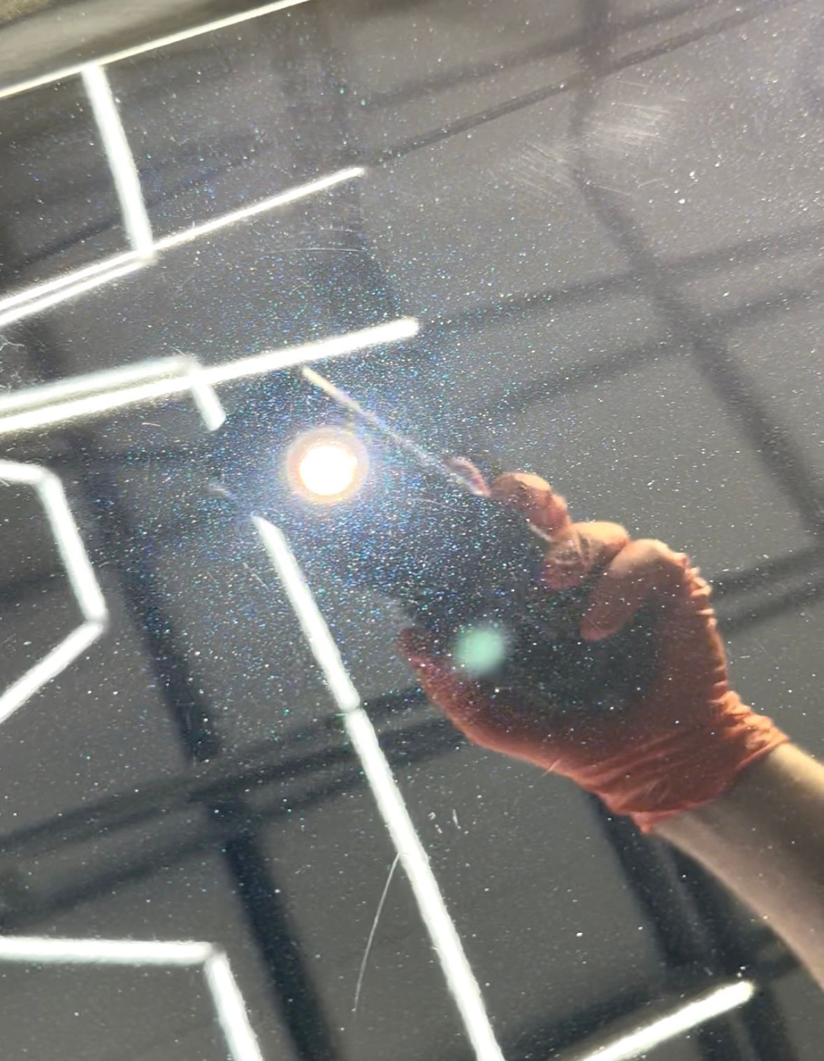 Hand holding a flashlight over a car surface, illuminating swirl marks and scratches. Bright light, orange glove.