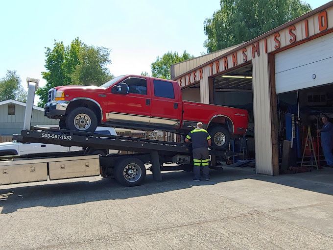 A red truck is sitting on top of a tow truck in front of a garage.