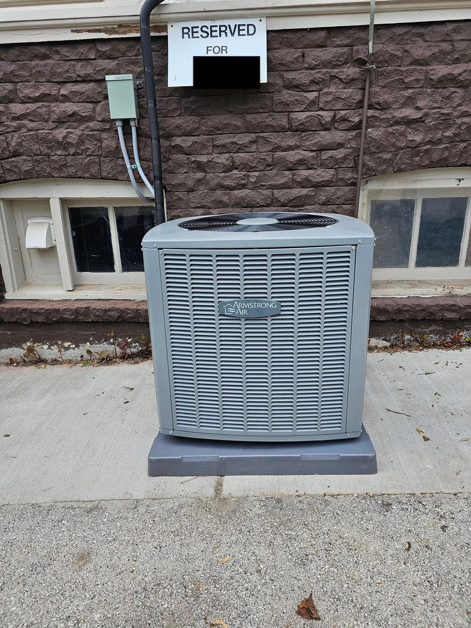 A gray air conditioner is sitting on the sidewalk in front of a brick building.