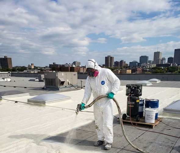 Man in protective suit spraying white foam on a rooftop; city buildings in background.