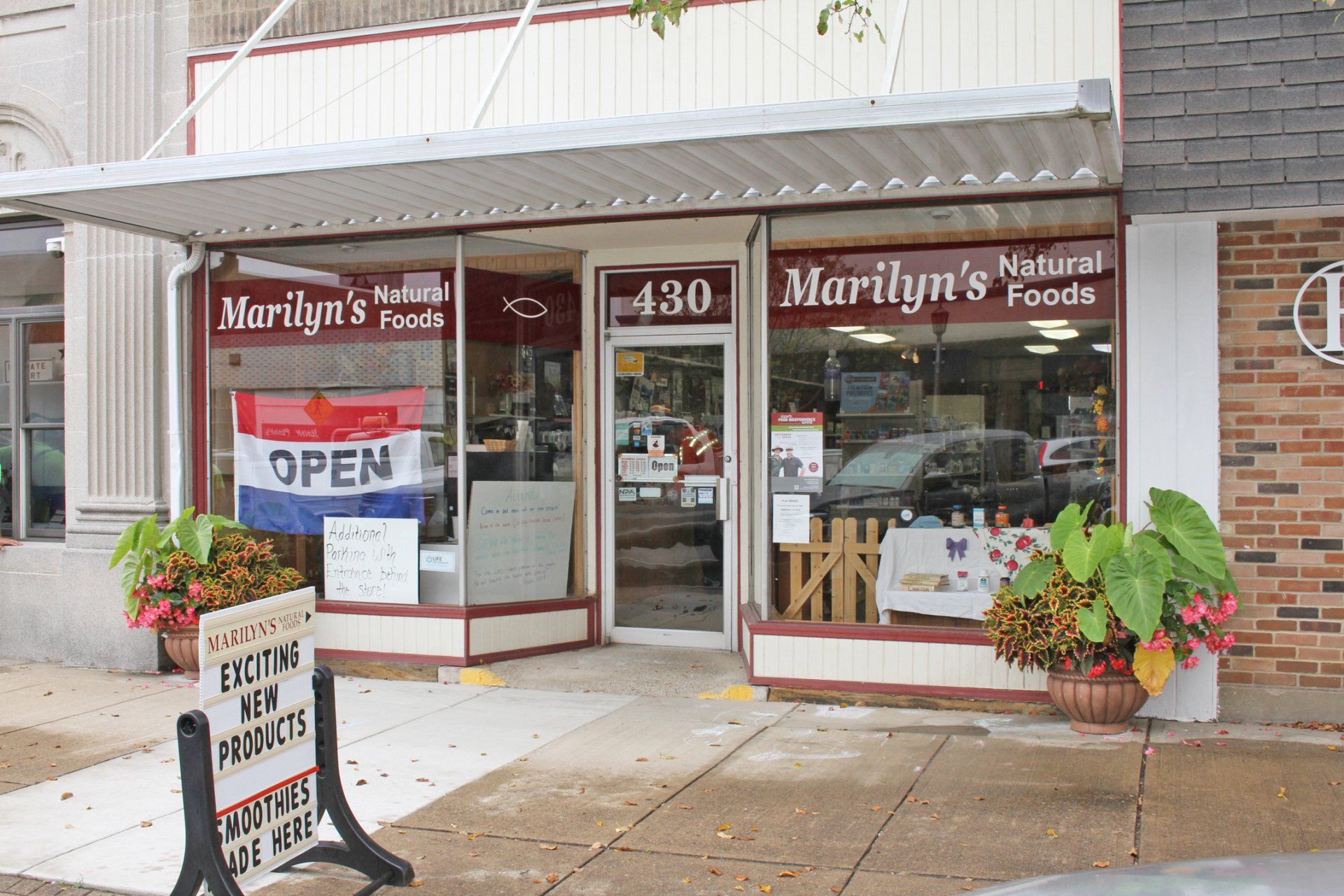 A storefront for Marilyn's Natural Foods at 430 with an open sign in the window, a sidewalk sign, and potted plants.